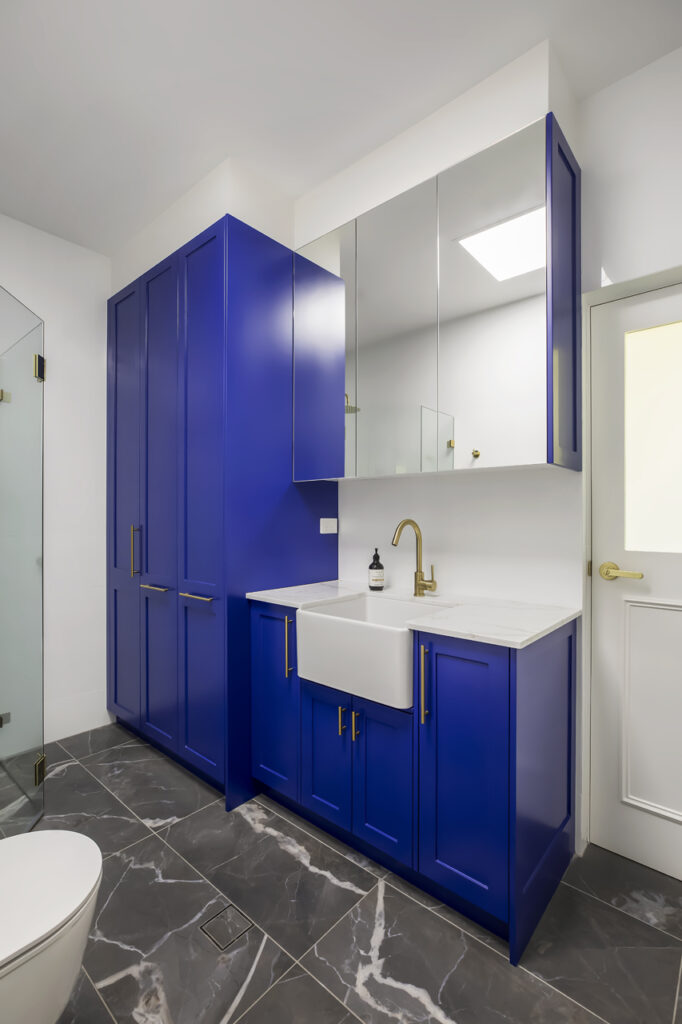 A bold and stylish bathroom renovation by HoverCo, featuring striking blue cabinetry, gold fixtures, a farmhouse sink, and elegant marble flooring for a modern luxury design.