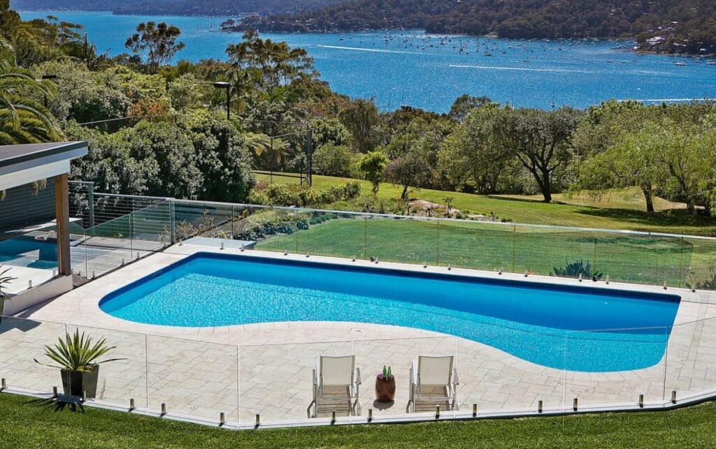 A luxury custom-designed swimming pool with glass fencing, installed by HoverCo, overlooking a stunning waterfront landscape with lush greenery.