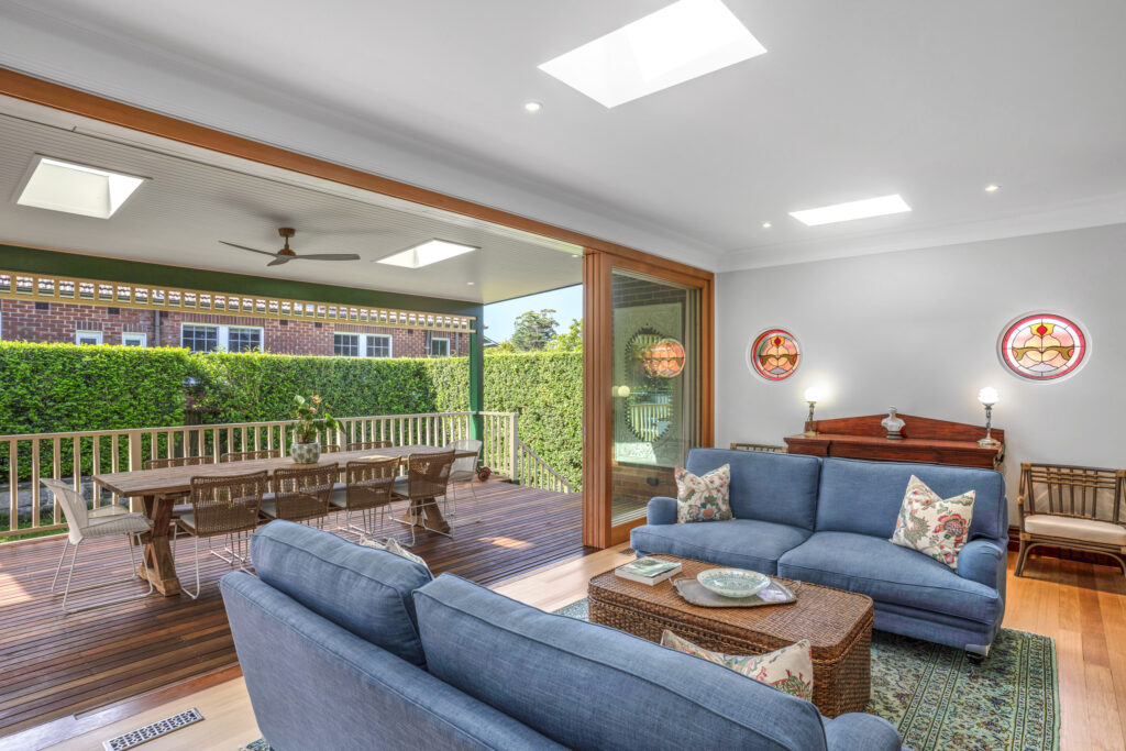 A beautifully designed indoor-outdoor living space by HoverCo, featuring a spacious timber deck, skylights, sliding glass doors, and a cozy lounge area with blue sofas and stained glass windows.