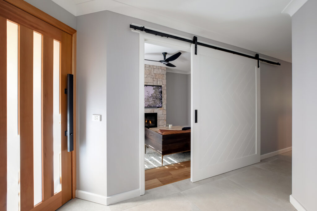 A modern entryway renovation by HoverCo, featuring a custom timber front door, a white barn-style sliding door with black hardware, and a cozy living space with a stone fireplace.