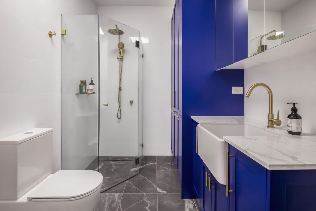 A stylish modern bathroom renovation by HoverCo, featuring a bold blue vanity, gold fixtures, a frameless glass shower, and sleek marble flooring for a sophisticated touch.