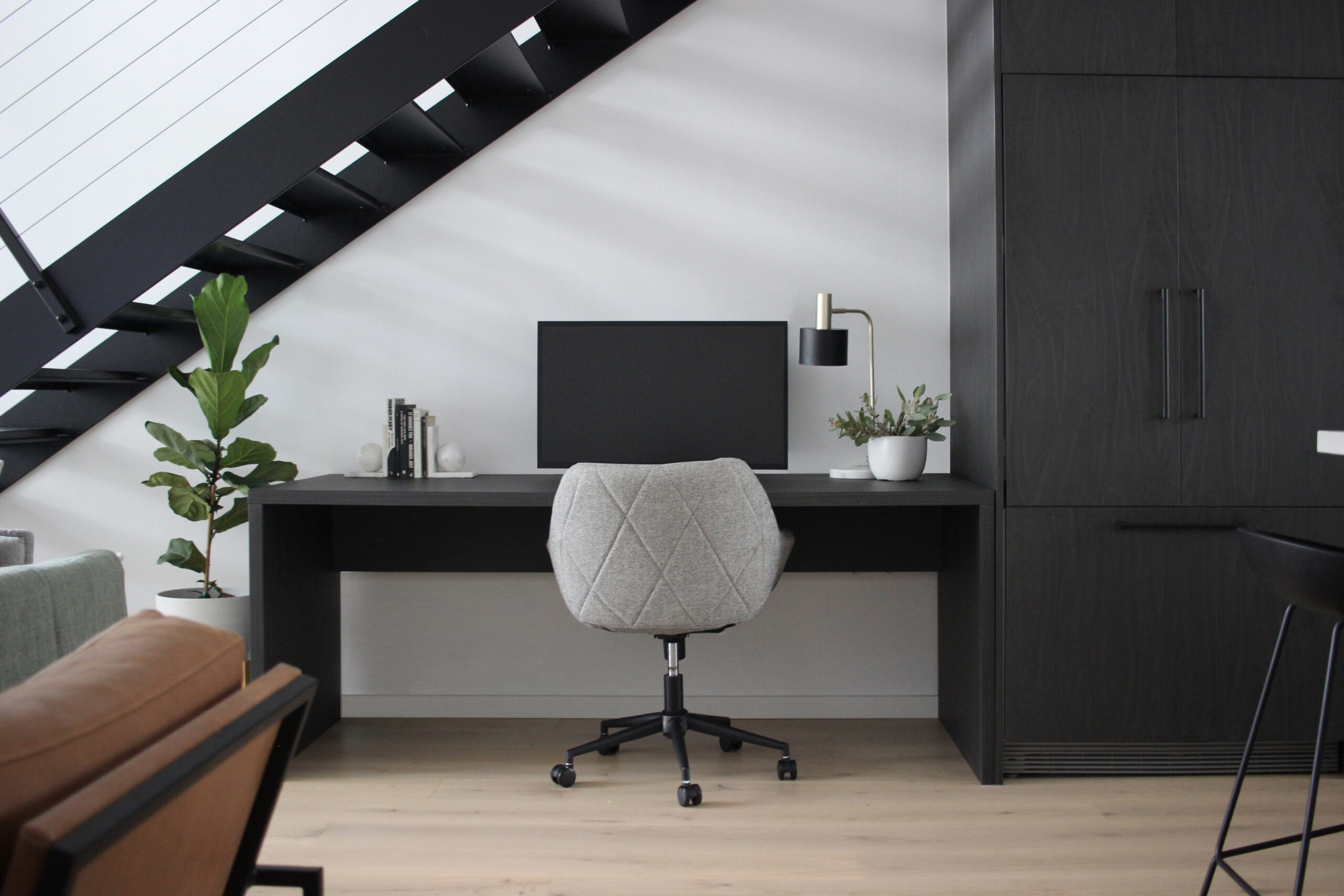A sleek modern home office designed by HoverCo, featuring custom black joinery, a minimalist desk setup, stylish lighting, and integrated storage under a staircase for a functional workspace