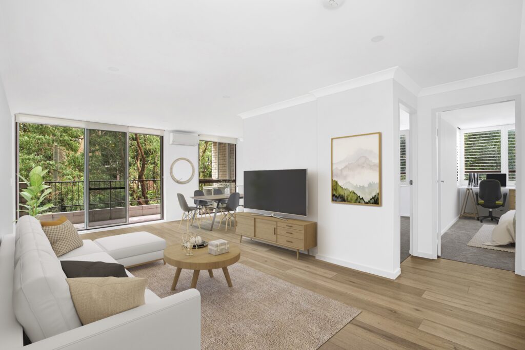 A bright and modern open-plan living room designed by HoverCo, featuring a white sectional sofa, wooden flooring, sliding glass doors leading to a balcony, and a cozy home office nook.