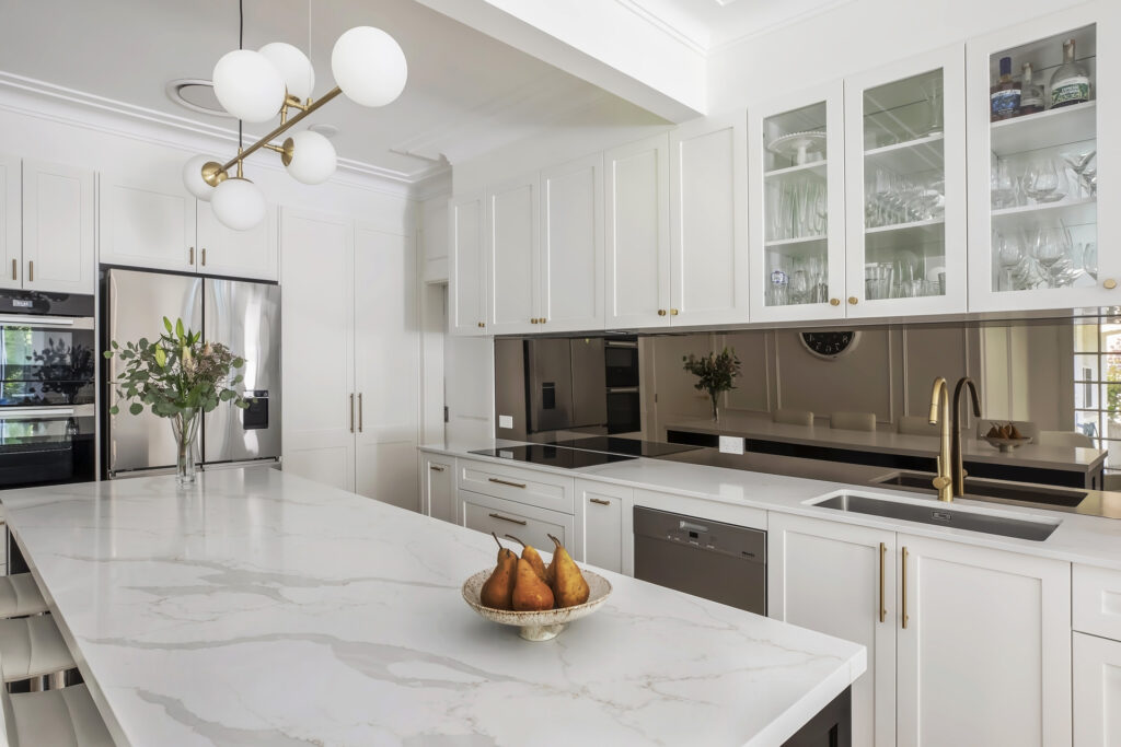 A modern white kitchen designed by HoverCo, showcasing a marble island, gold hardware, glass-front cabinets, and premium stainless steel appliances for a timeless aesthetic.