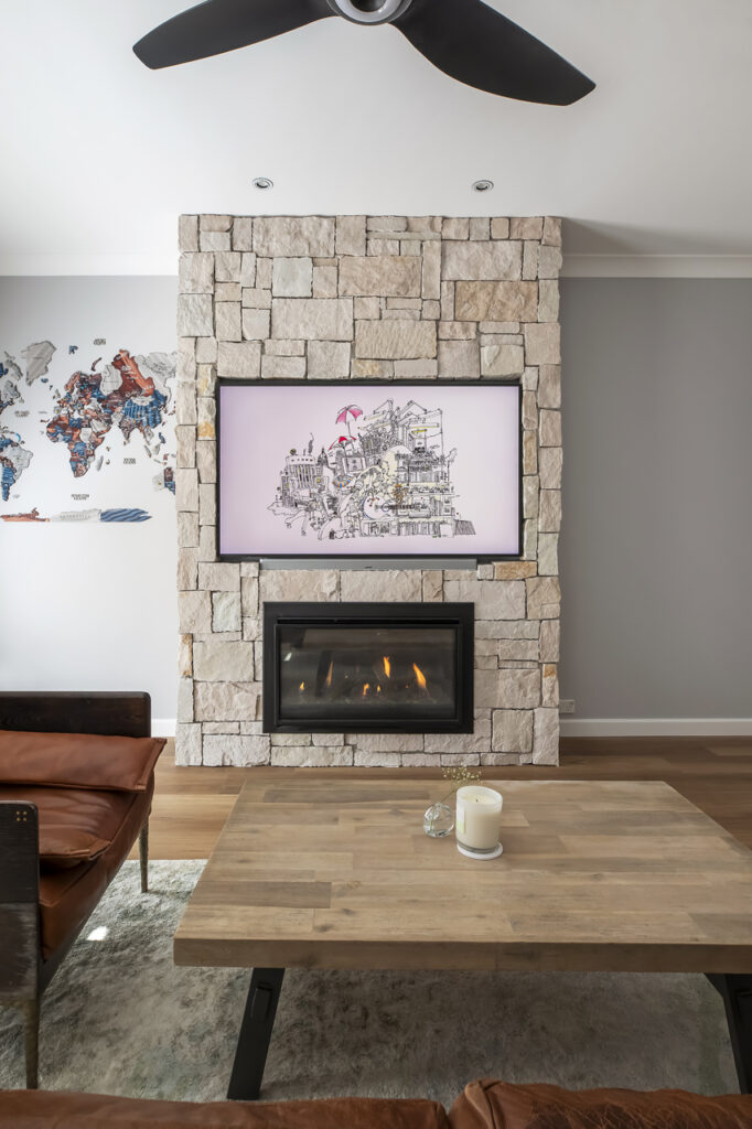 A cozy living room renovation by HoverCo, featuring a rustic stone fireplace, a built-in TV display, warm leather seating, and a wooden coffee table for a modern yet inviting atmosphere.