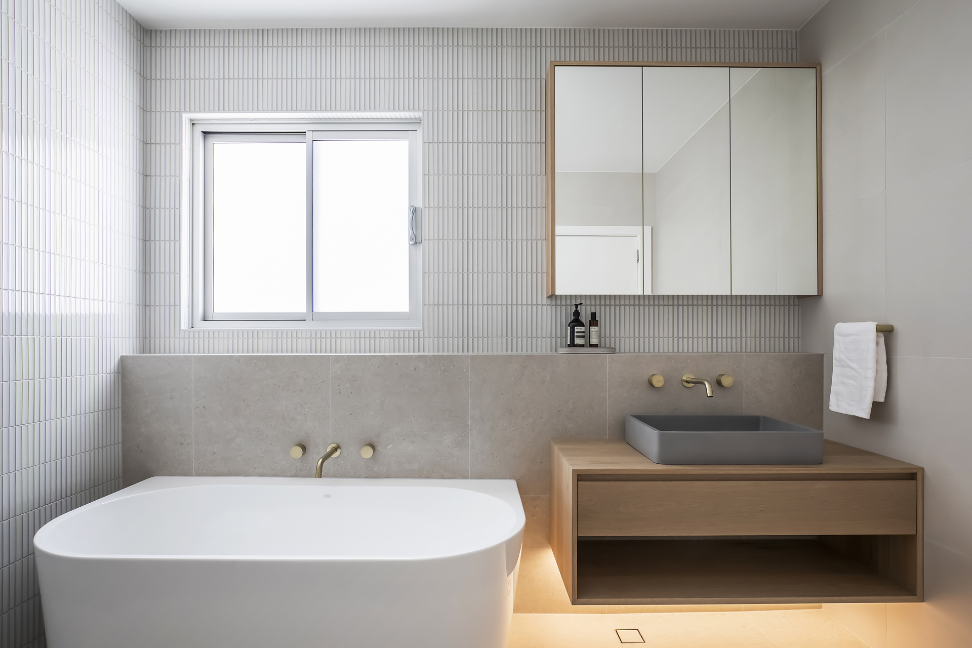 A sleek and modern bathroom renovation by HoverCo, featuring a freestanding bathtub, floating wooden vanity, gold fixtures, and soft ambient lighting for a contemporary spa-like feel.